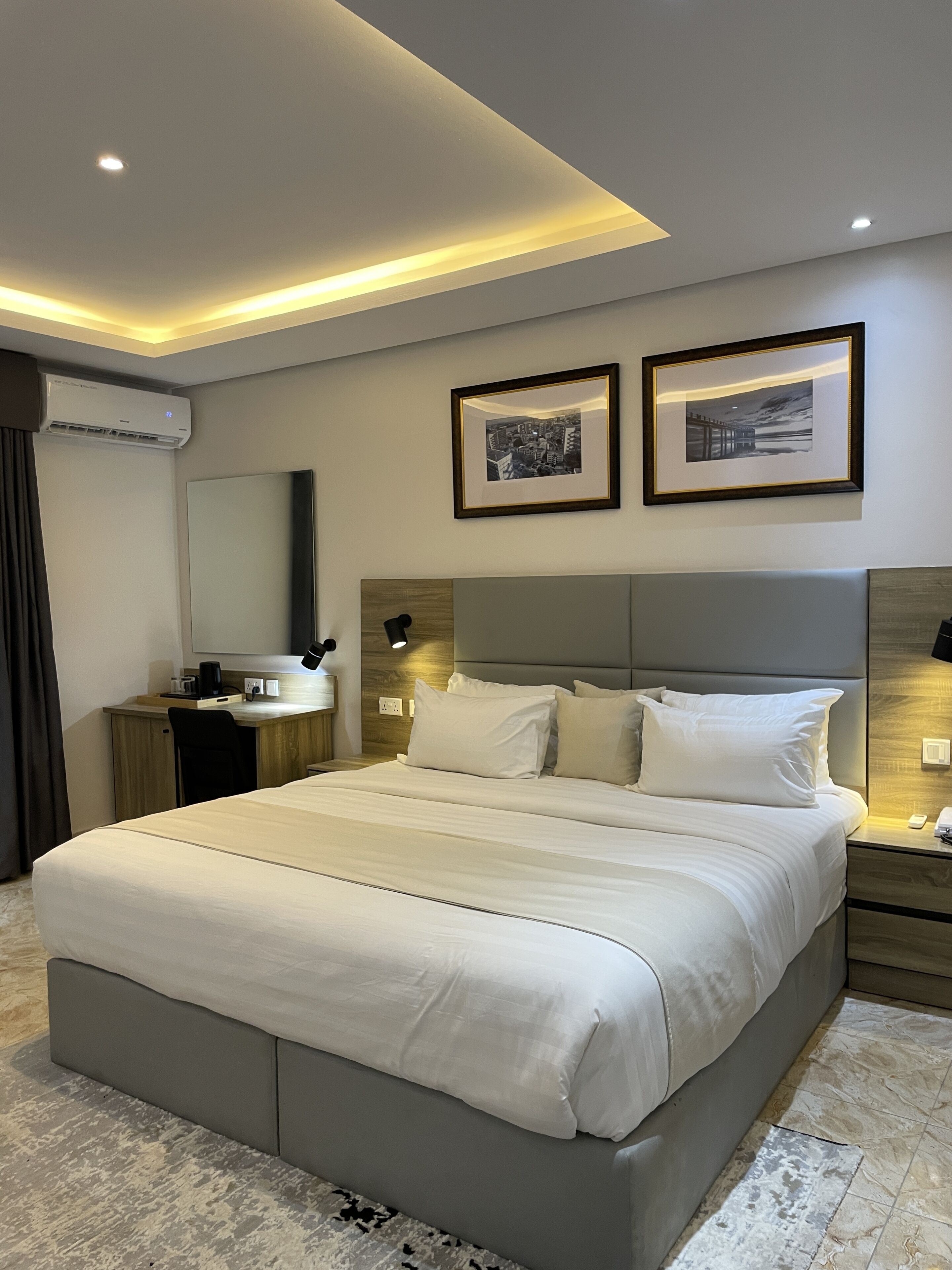 Una habitación de hotel moderna con una cama grande, mesitas de noche, un escritorio con silla, un equipo de aire acondicionado y cuadros en la pared.