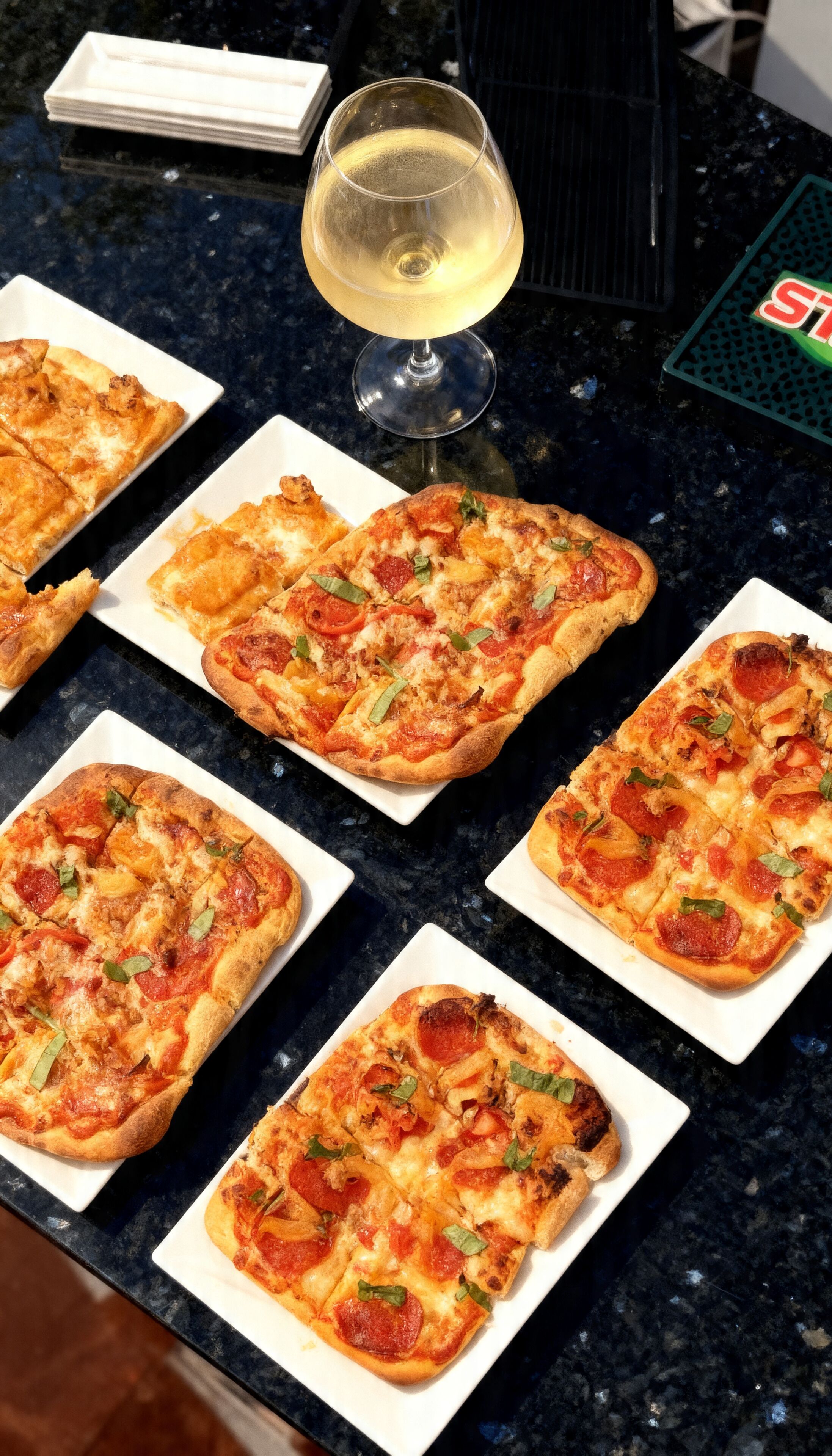 Une table sur laquelle se trouvent plusieurs pizzas carrées garnies de pepperoni et de feuilles vertes, un verre de vin blanc et un plateau vert orné d’un logo rouge.