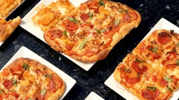 Une table sur laquelle se trouvent plusieurs pizzas carrées garnies de pepperoni et de feuilles vertes, un verre de vin blanc et un plateau vert orné d’un logo rouge.
