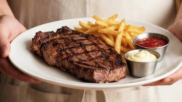 A person holding a plate with steak, fries, and condiments, and a salad on a table.