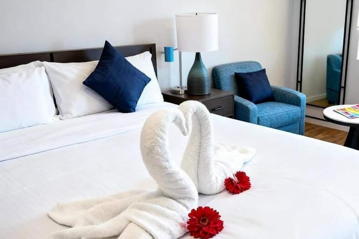 A hotel room with a neatly made bed, a blue armchair, a small table with a lamp, and a mirror on the wall.