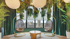 A room with a round wooden table, two wicker chairs, hanging plants, and a view of the city through large windows.