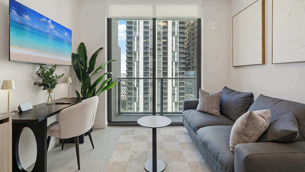 A modern living room with a sofa, a round table, and a desk with a chair. There is a large window with a view of high-rise buildings.