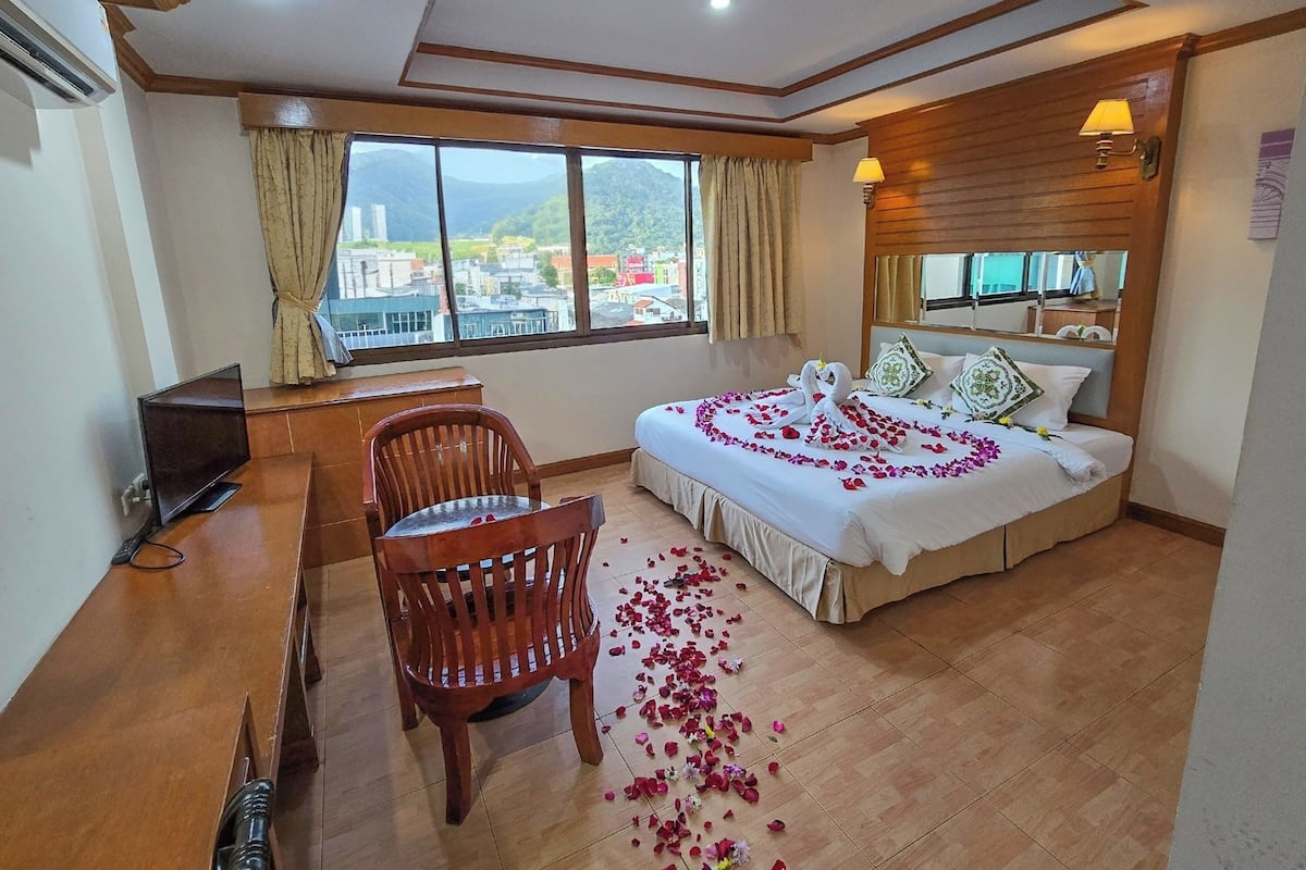 A hotel room with a bed, a desk, a chair, a TV, and a large window with a view of buildings and hills.