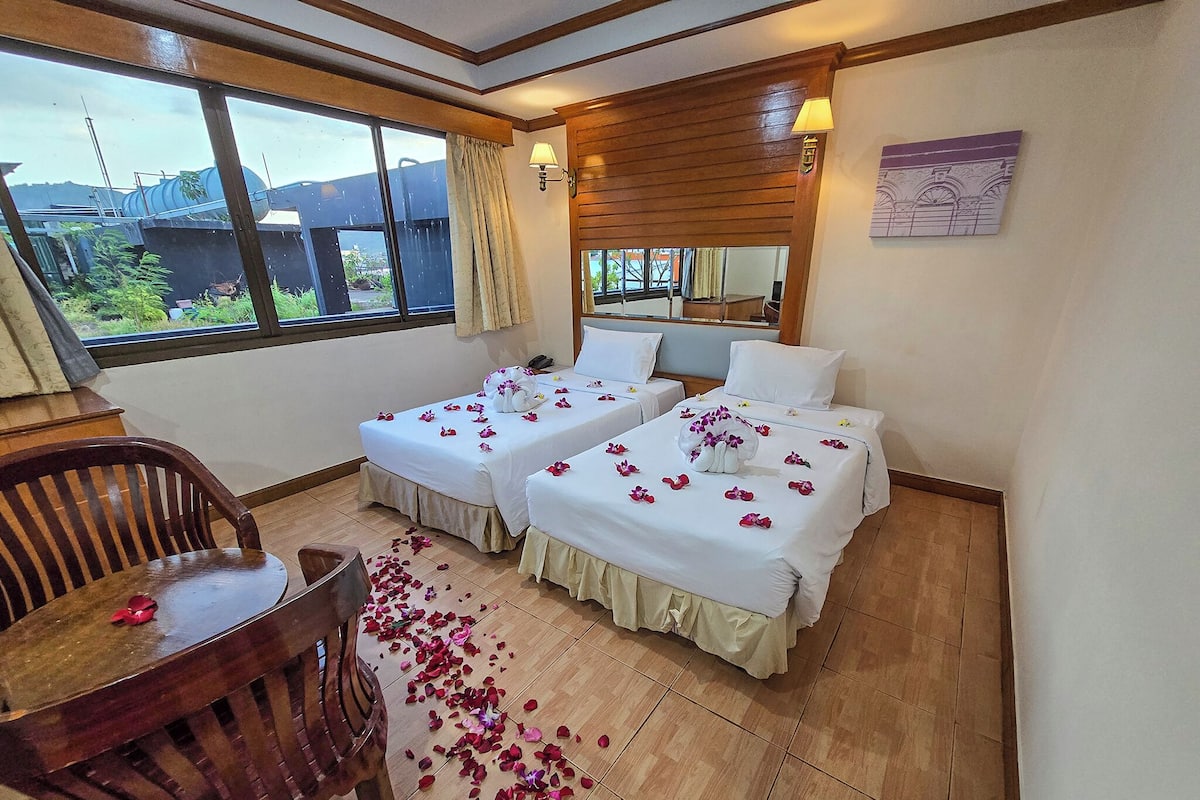 A hotel room with two beds, a wooden chair, a table, and a large window with a view of greenery.
