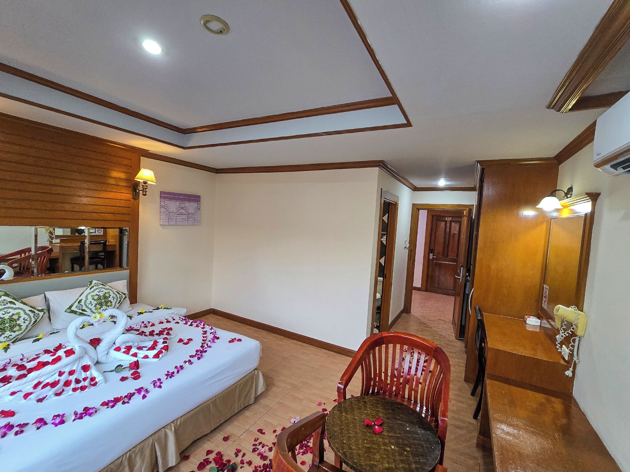 A hotel room with a bed, a chair, a table, a mirror, and a hallway leading to another room.