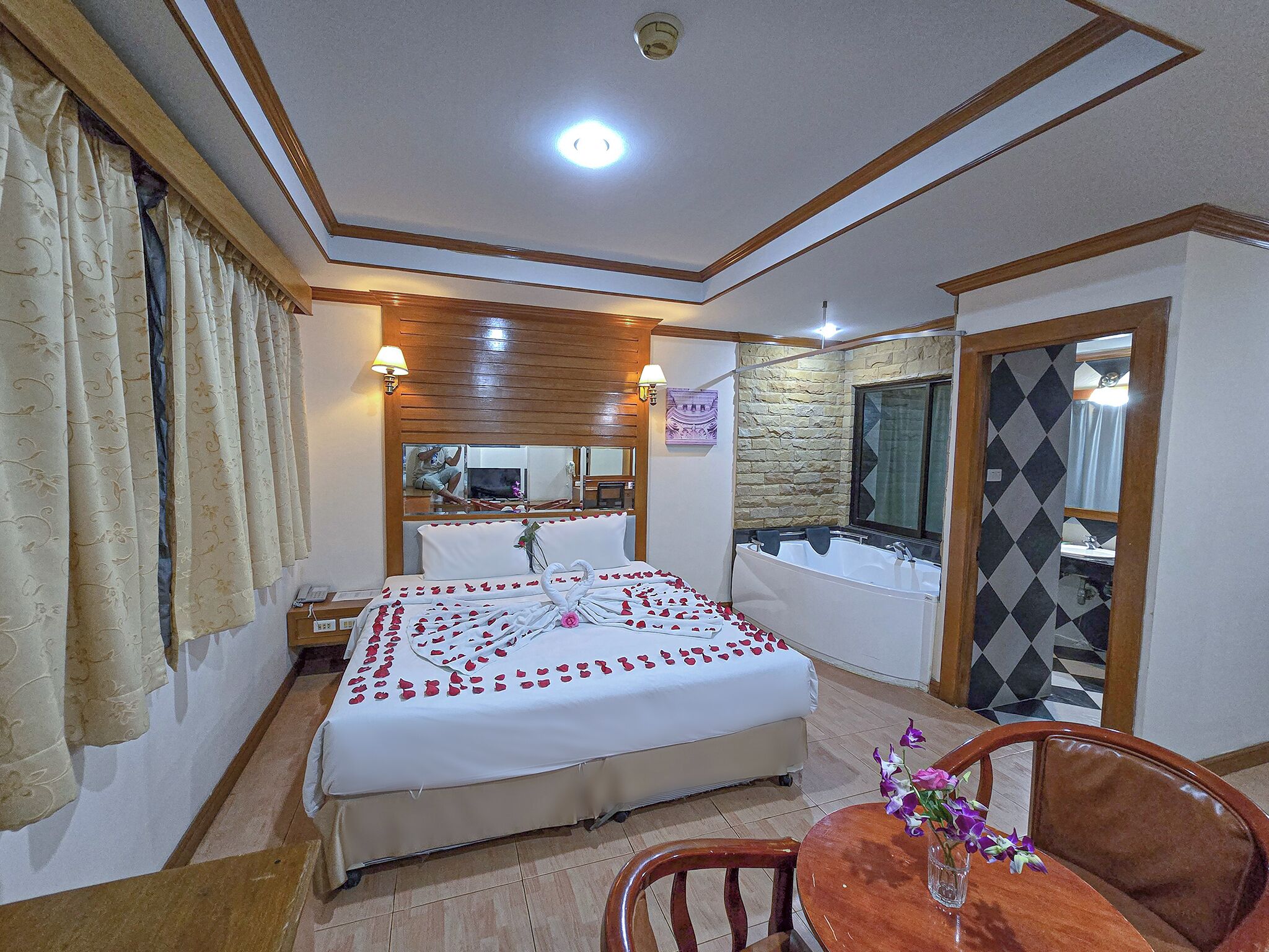A hotel room with a bed, a bathroom with a bathtub, a dining table with chairs, and a mirror.