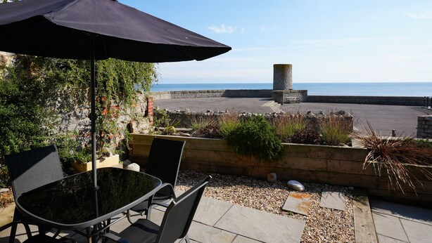 An outdoor patio with a table and chairs, a black umbrella, and a view of the sea and a stone structure. - Blue Horizons seaside flat in town centre (Lyme Regis)