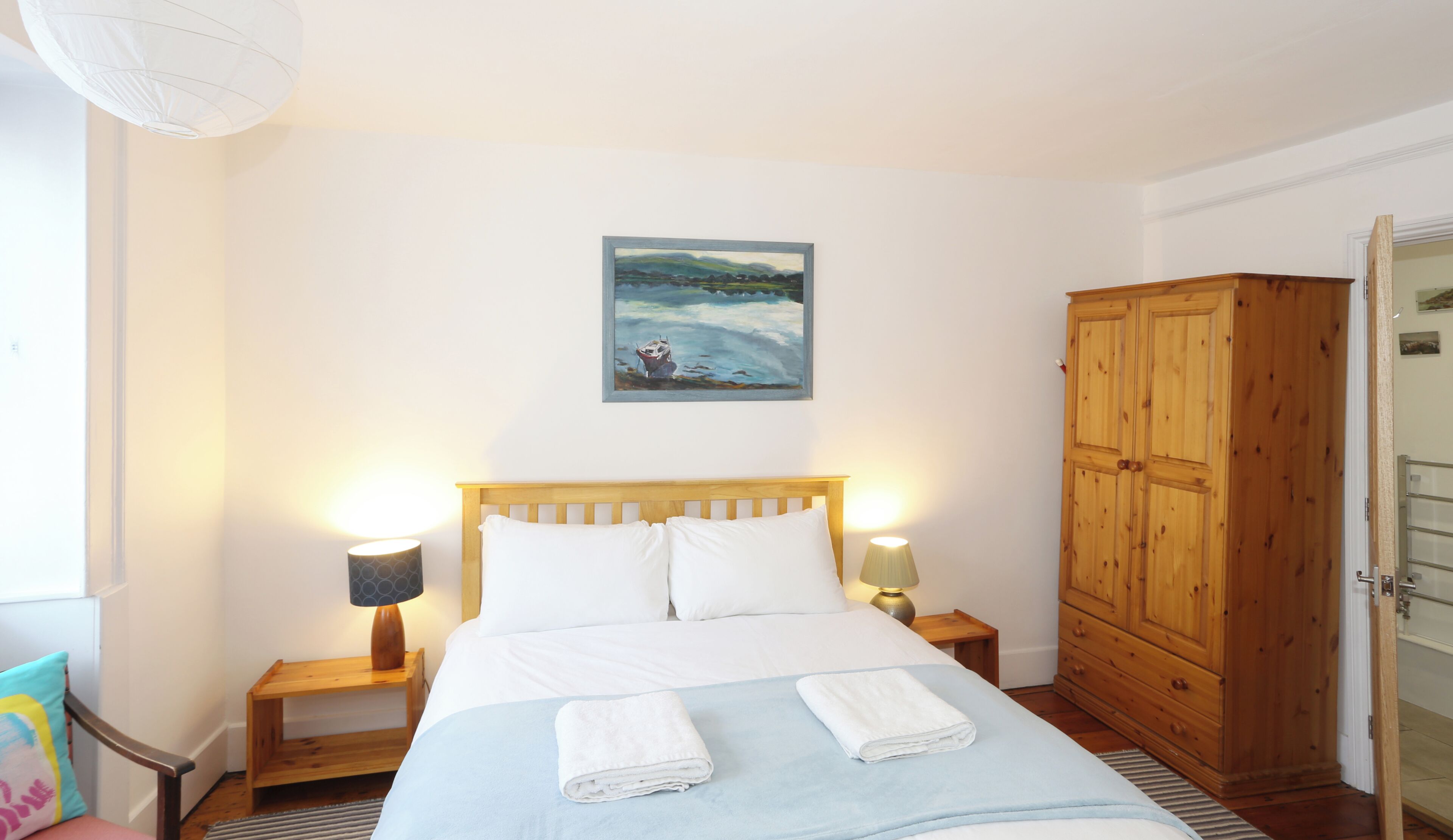 A bedroom with a bed, two bedside lamps, a wooden wardrobe, and a painting on the wall.
