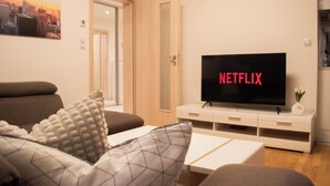 Ein Wohnzimmer mit einem Fernseher, der 'NETFLIX' zeigt, einer Couch, einem Couchtisch und einer Tür zu einem anderen Raum.