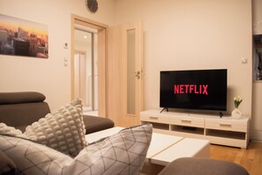 Una sala de estar con una televisión mostrando 'NETFLIX', un sofá, una mesa de centro y una puerta que da a otra habitación.