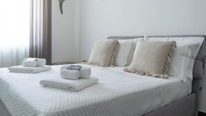 Ein ordentlich bezogenes Bett mit weißer Bettwäsche und zusammengerollten Handtüchern, eine Wandleuchte und ein dekoratives Wandbild.