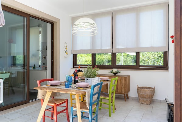 Un coin repas avec une table dressée pour quatre personnes, entouré de chaises colorées, d’un luminaire suspendu et offrant une vue sur la verdure à travers les fenêtres.