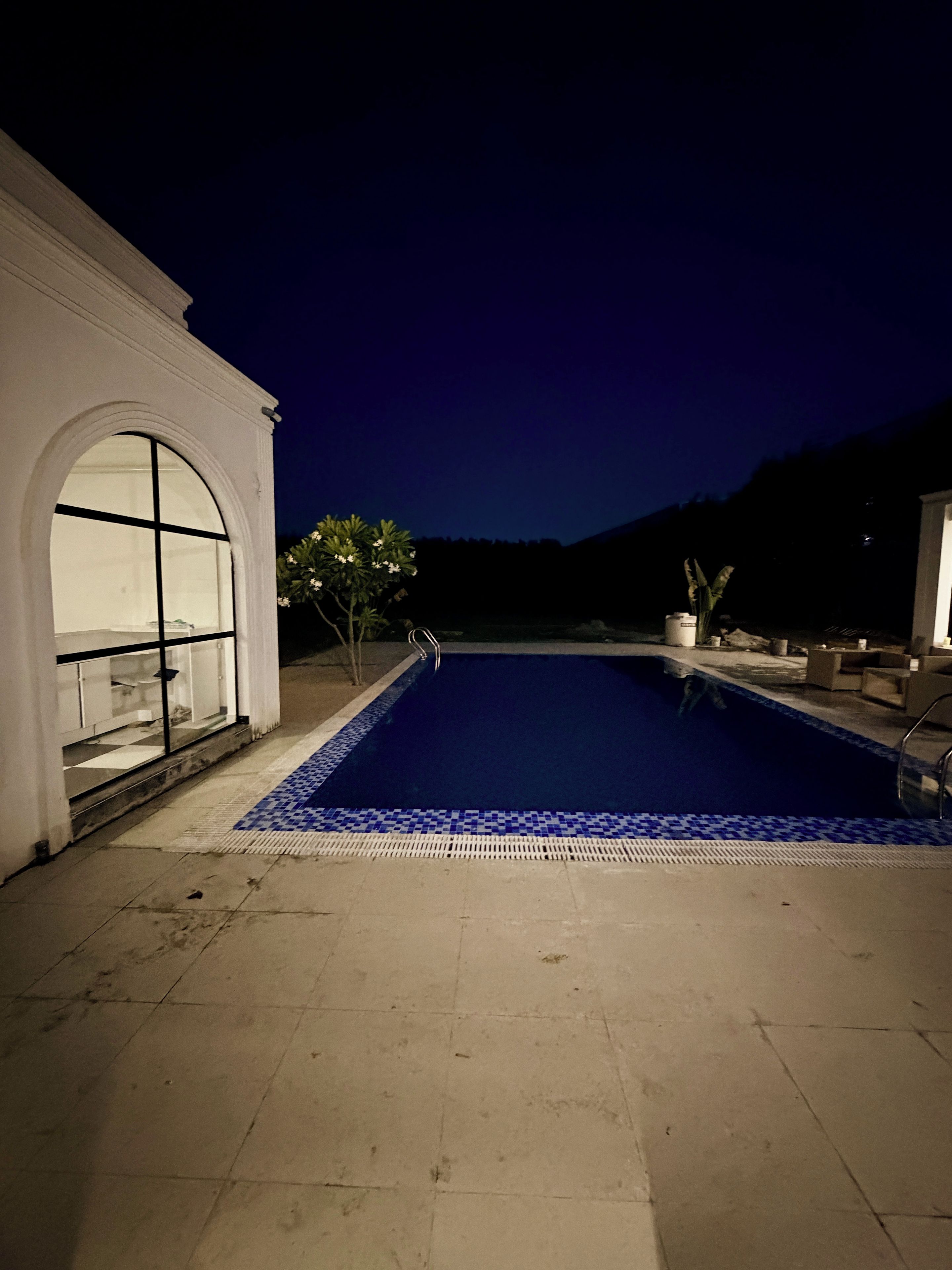 Una piscina con borde de azulejos, un edificio blanco con un amplio ventanal arqueado y mobiliario exterior.