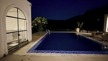 Una piscina con borde de azulejos, un edificio blanco con un amplio ventanal arqueado y mobiliario exterior.