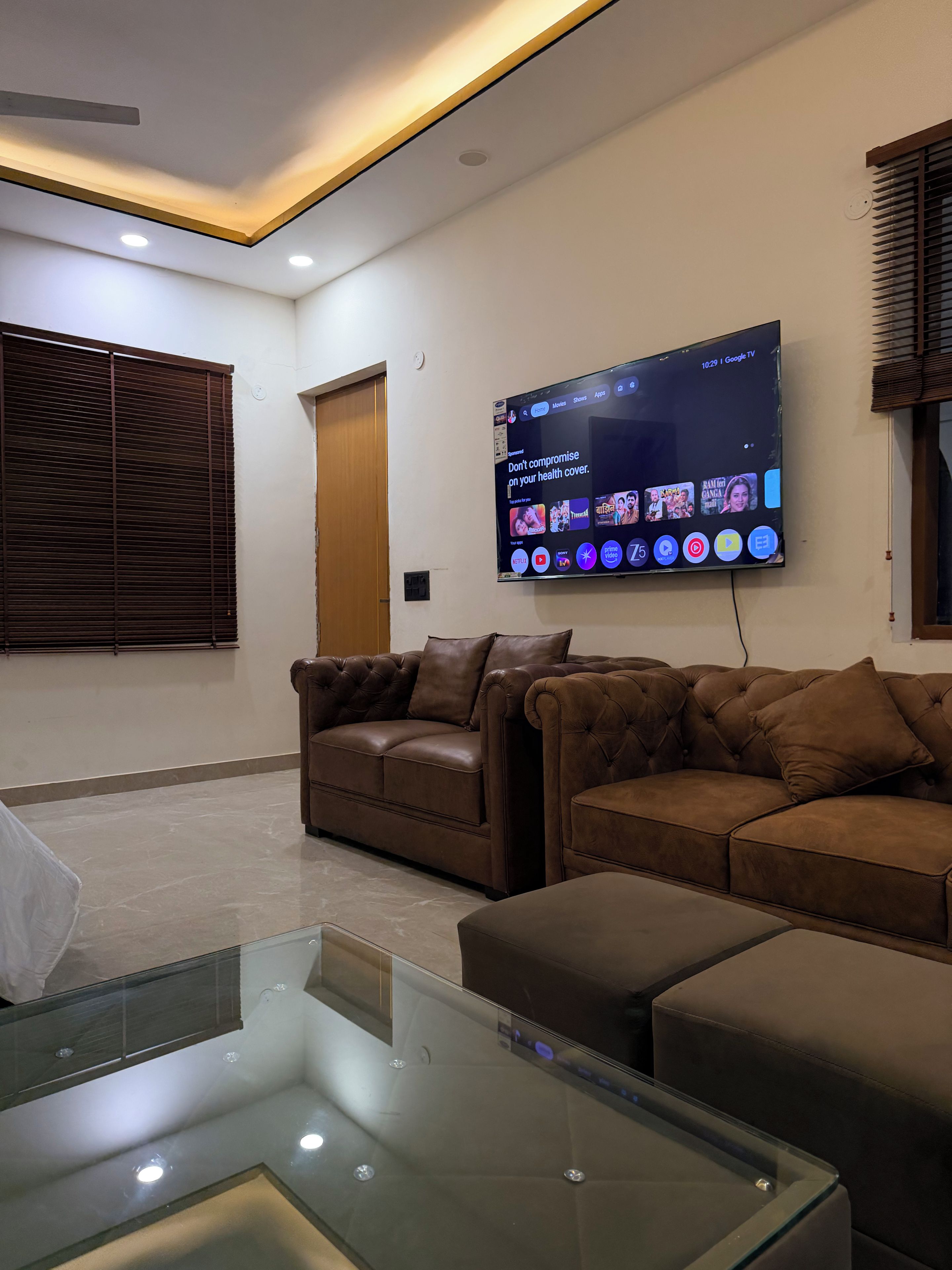 A modern living room with a flat-screen TV mounted on the wall, two leather sofas, a glass coffee table, and recessed ceiling lighting.