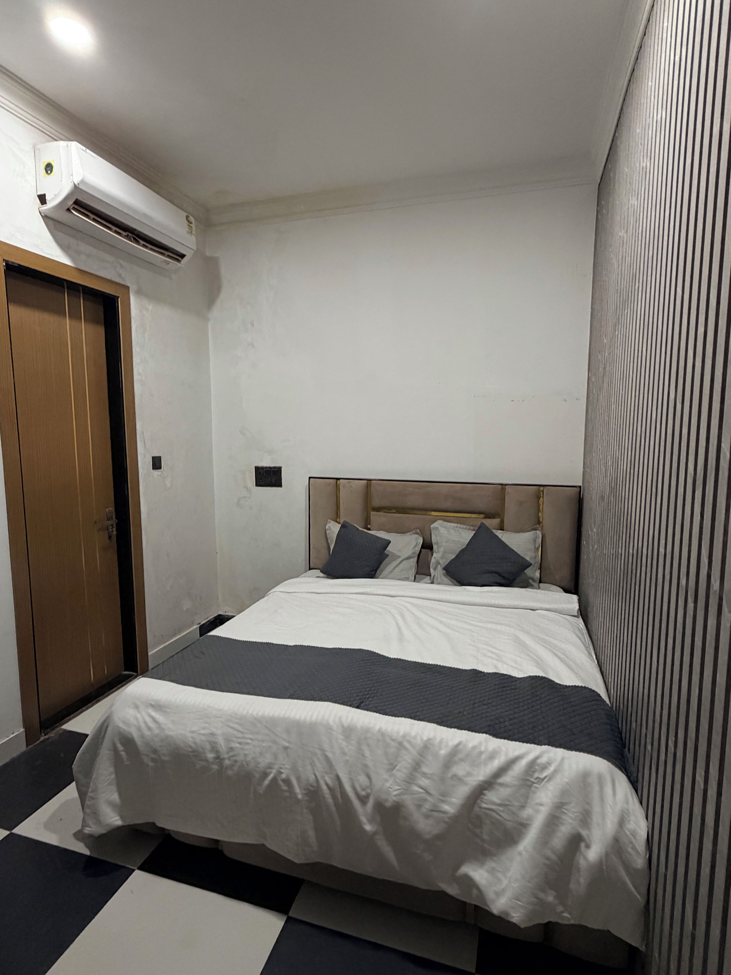 Un dormitorio pequeño y ordenado con una cama individual, una puerta de madera y un equipo de aire acondicionado instalado en la pared.