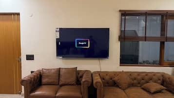 A room with two brown leather sofas, a flat-screen TV mounted on the wall, and a window with blinds.