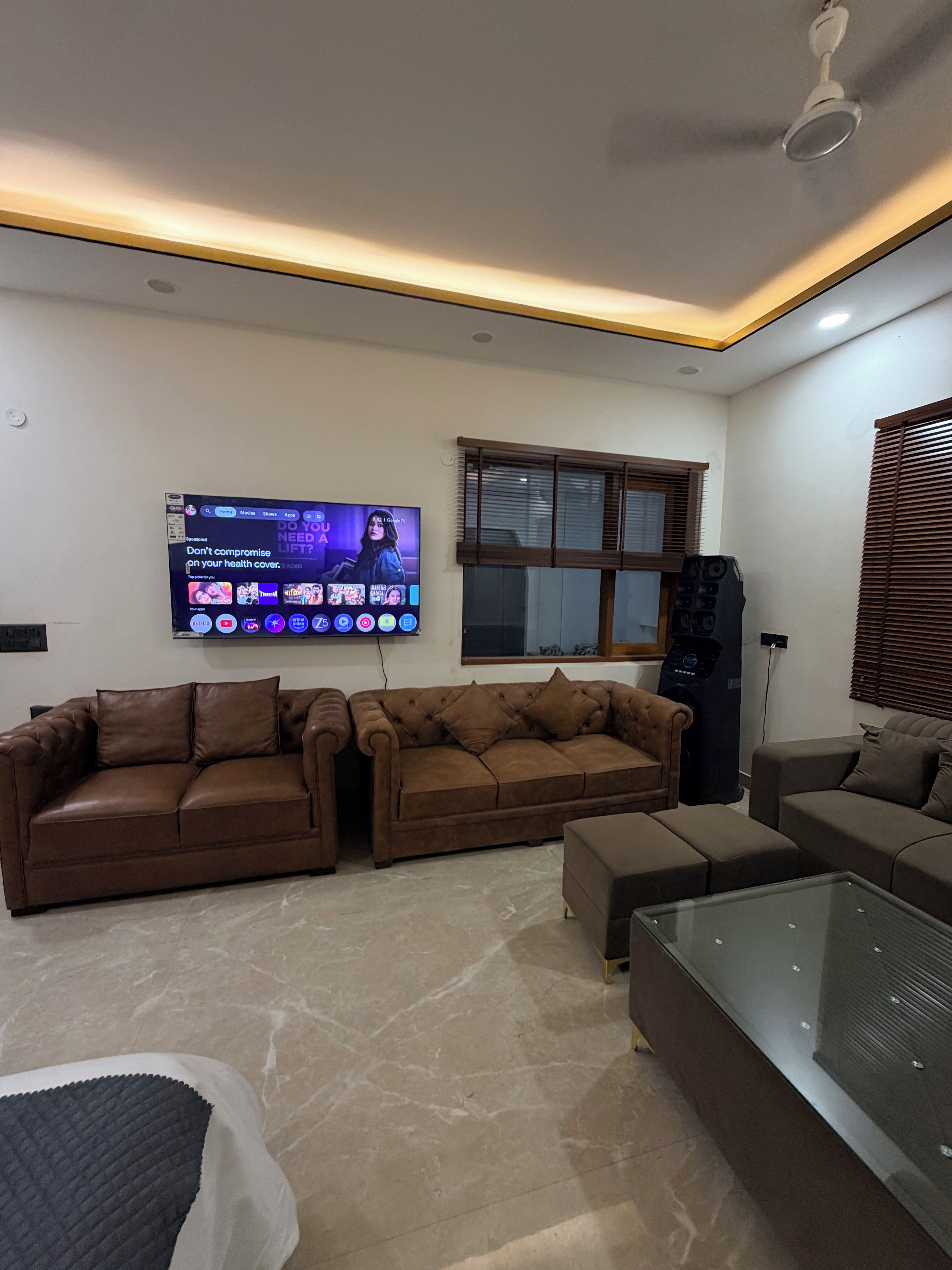 Una sala moderna con un televisor de pantalla plana montado en la pared, dos sofás de cuero marrón, una mesa de centro de vidrio y un ventilador de techo.
