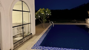A swimming pool with a tiled edge, a white building with a large arched window, and a small tree with white flowers. - 2BR Villa with Pool, BBQ & Music at GoBravo Grandeur Gurgaon (Gurugram)