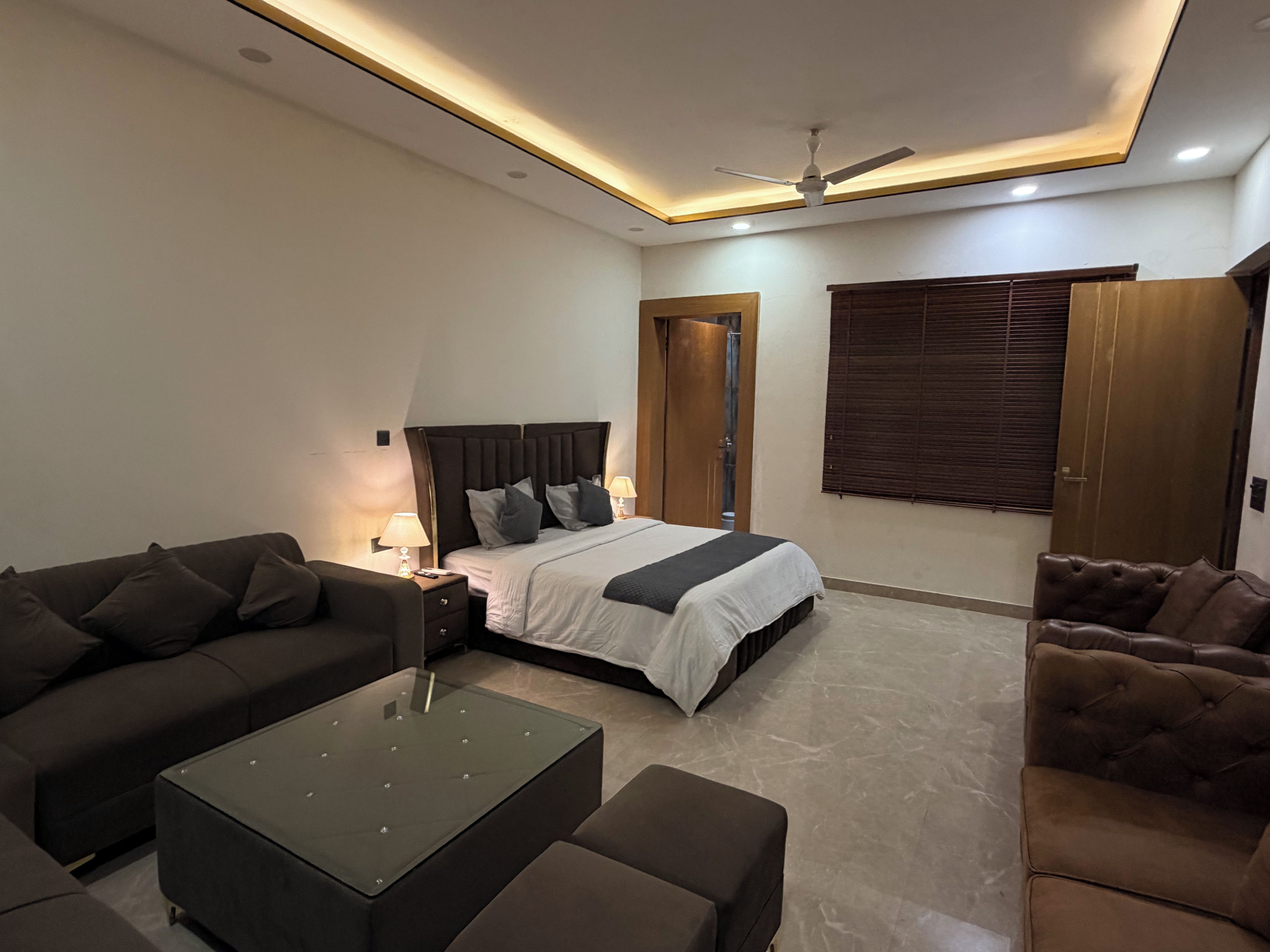 A modern hotel room with a bed, two sofas, a glass coffee table, and a ceiling fan.