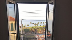 Ein Schlafzimmer mit großem Fenster, einem Bett und Blick auf eine Küstenlandschaft mit Gebäuden und Palmen.