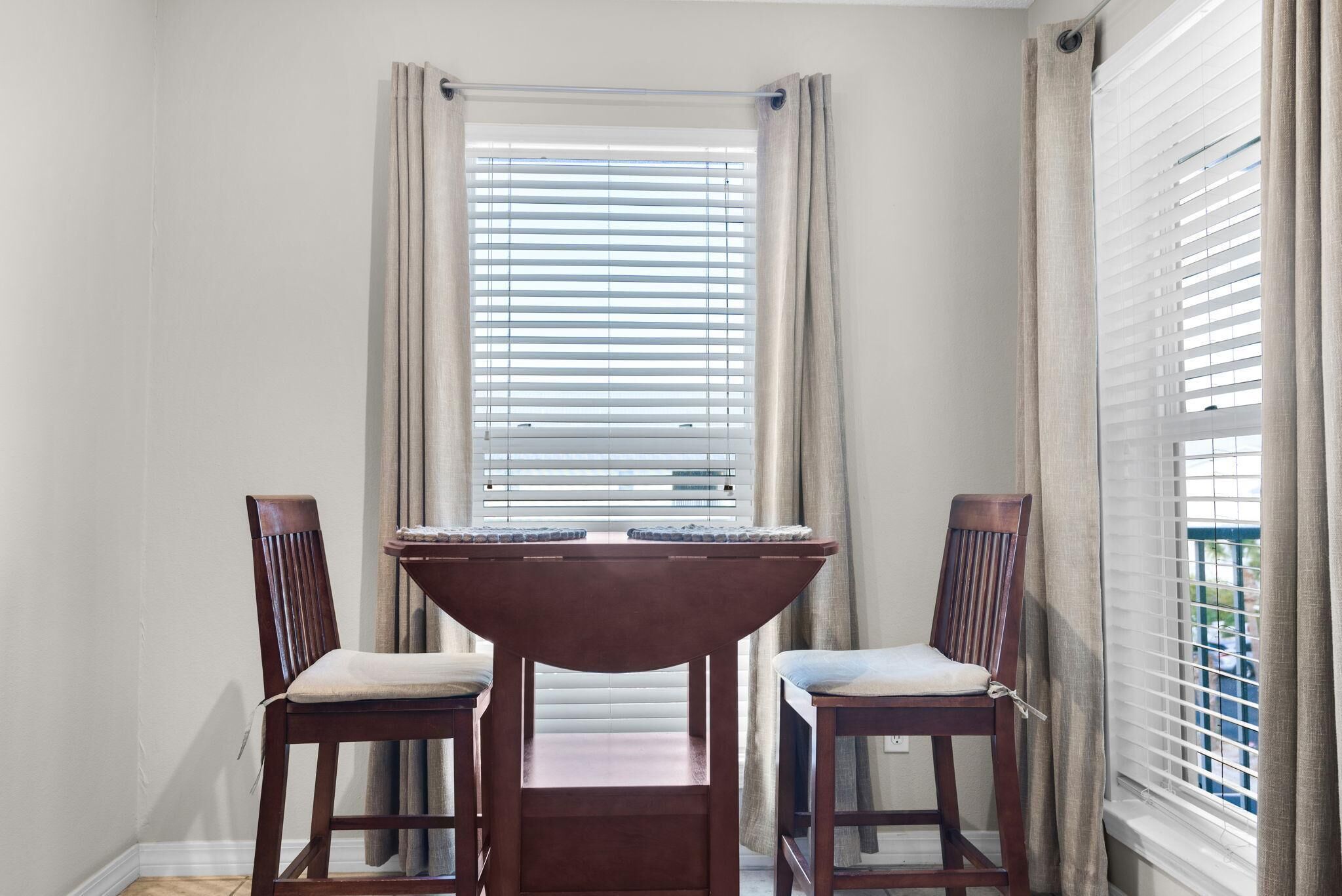 A dining area with a round table and two chairs, a window with blinds, and a view of the outside.
