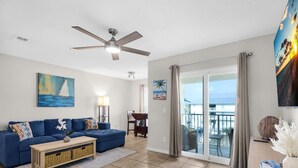 A living room with a blue sofa, a wooden coffee table, a ceiling fan, and a view through a sliding glass door to the outside. - Gulf Island 532 (Navarre)