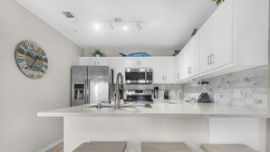 A modern kitchen with white cabinetry, stainless steel appliances, a clock, and a 'Welcome' sign. - Gulf Island 532 (Navarre)