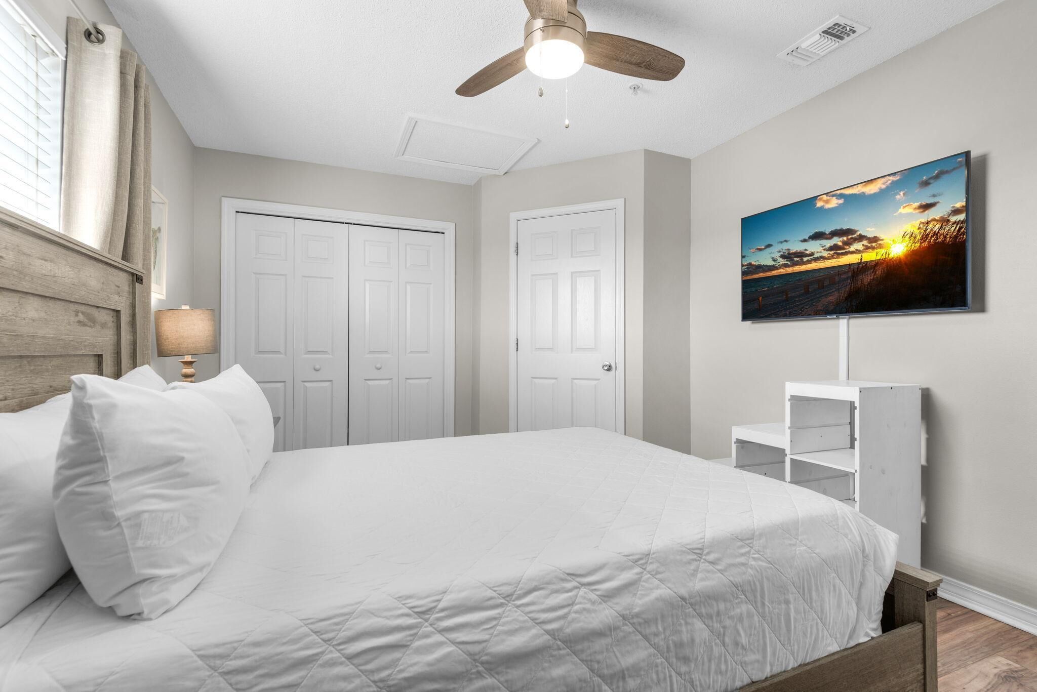 A bedroom with a bed, a ceiling fan, a TV on the wall, and a window with curtains.