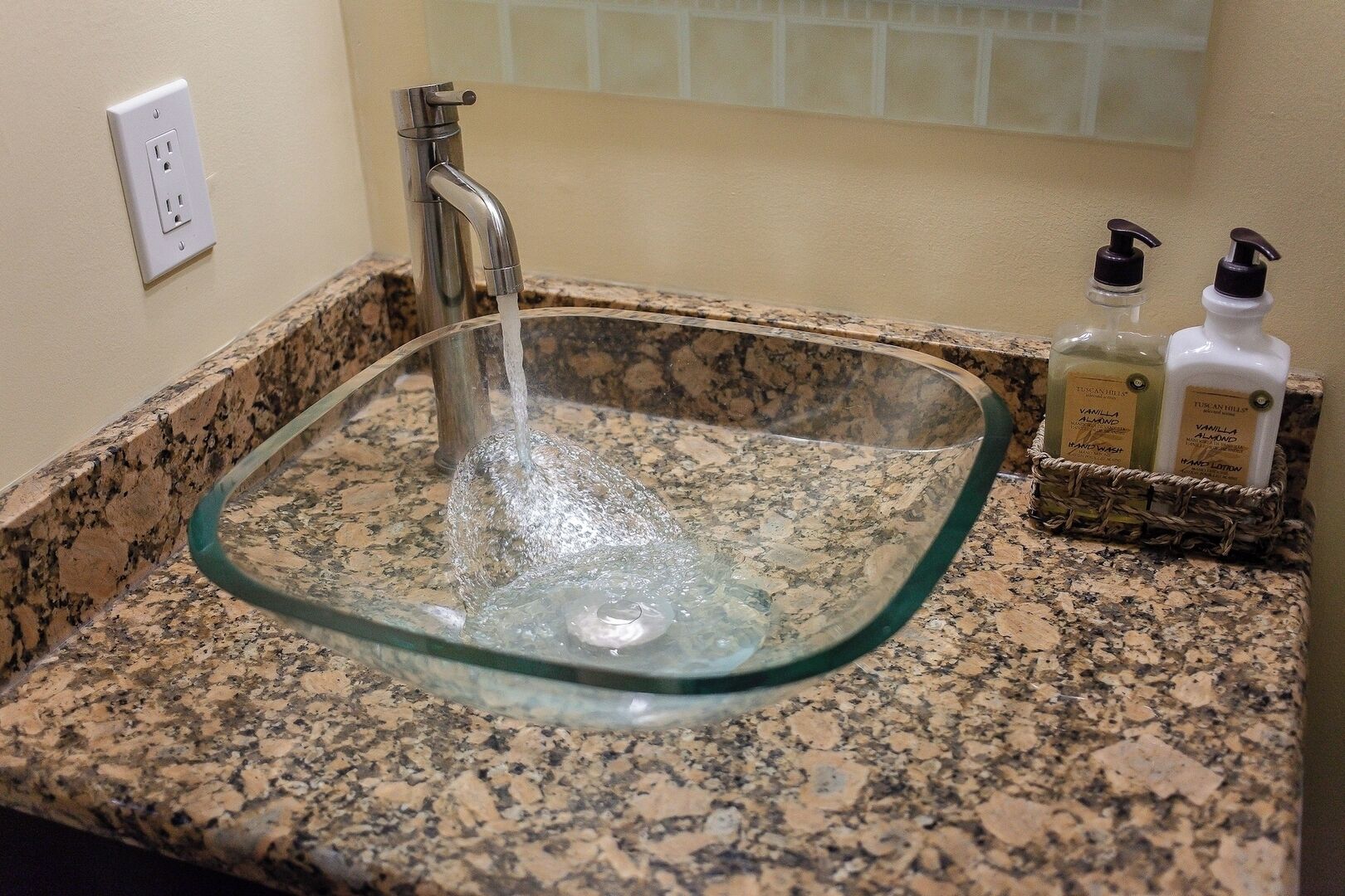 A glass sink with running water, a granite countertop, and two bottles of hand lotion.