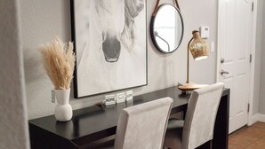 Een moderne eethoek met een donker houten tafel, twee stoelen, een ingelijst paardenportret, een vaas met gedroogde planten, een ronde spiegel en een lamp.