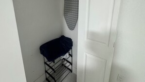 Uma pequena área de lavanderia com um varal de metal, uma tábua de passar roupa pendurada e uma porta branca.