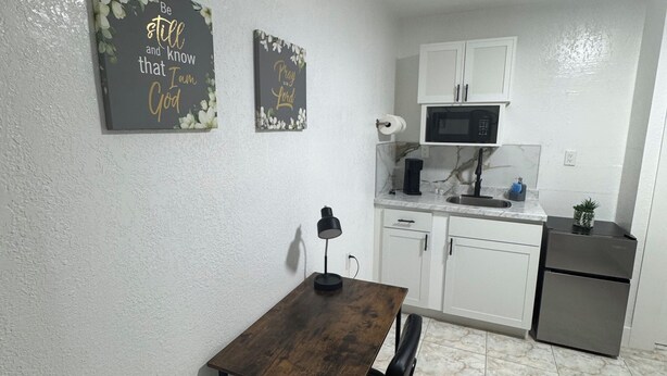 Cozinha compacta com armários brancos, micro-ondas e pequena geladeira. Há uma mesa de jantar com uma cadeira e duas placas na parede.