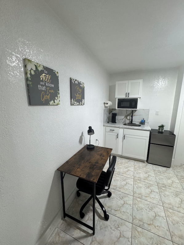 Una cocina compacta con gabinetes blancos, un microondas y un refrigerador pequeño. Hay una mesa de comedor con una silla y dos letreros en la pared.
