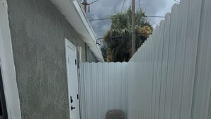 Um caminho de concreto estreito leva a uma porta branca, ladeado por uma cerca branca e um edifício com parede texturizada.
