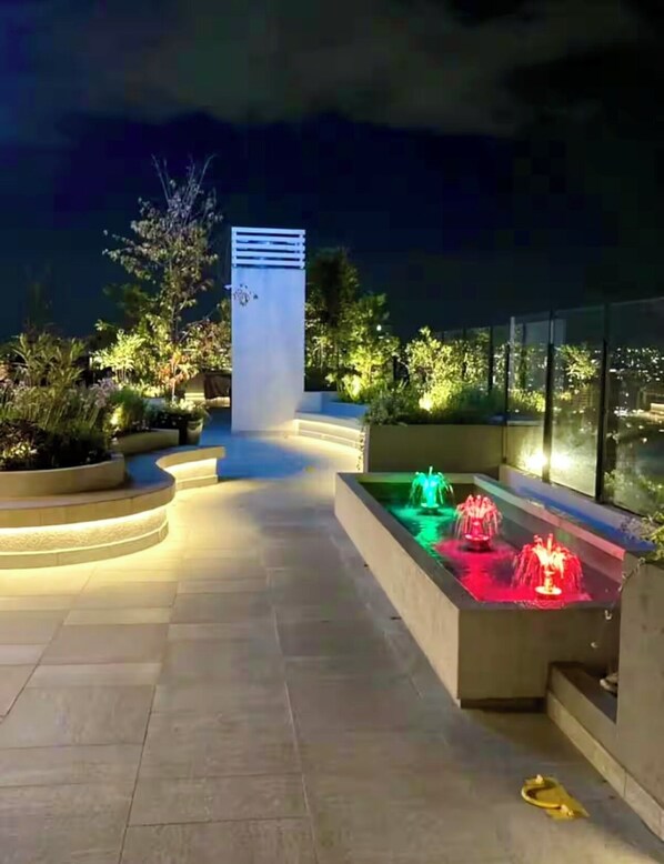 Spazio esterno con fontana, un'alta colonna e vista sulla città di notte.