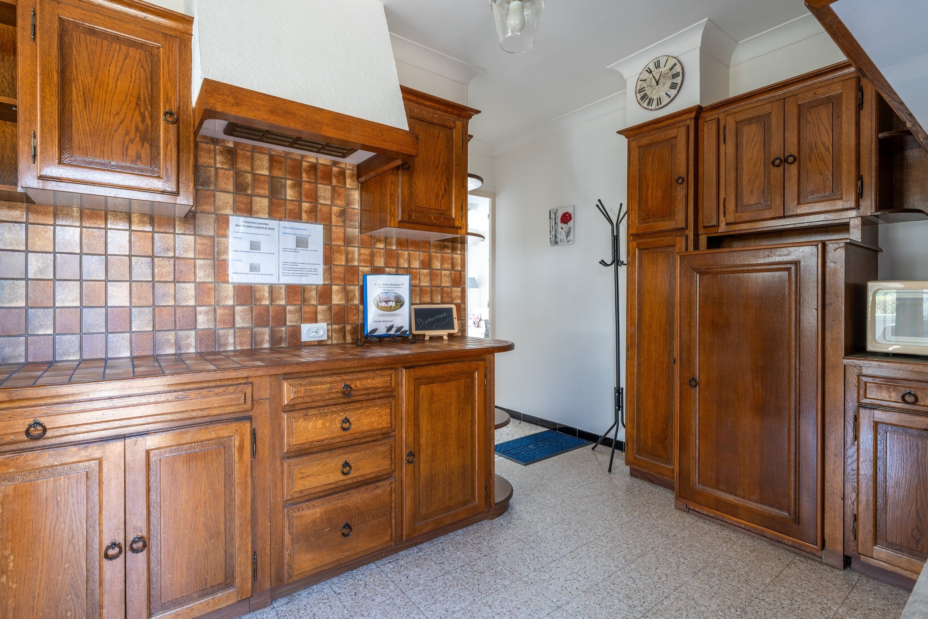 A kitchen with wooden cabinets, a tiled backsplash, a clock on the wall, and a microwave.