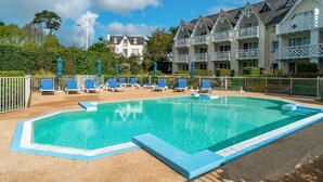 Ein Schwimmbecken mit Liegestühlen, einem Zaun und einem Gebäude mit Balkonen im Hintergrund.