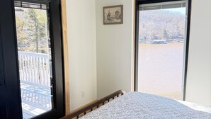Un dormitorio con cama, dos ventanas, una puerta, un pequeño cuadro enmarco y vistas a una terraza y árboles.
