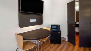 A compact room with a flat-screen TV mounted on the wall, a small black refrigerator, a wooden dining table with two chairs, and a door leading to another room. - Vagabond Inn - Santa Barbara (Santa Barbara)