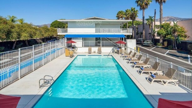 A pool with lounge chairs, a fenced area, and a building with a balcony in the background. - Vagabond Inn - Santa Barbara (Santa Barbara)