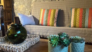 Una habitación acogedora con pared de piedra, suelo de madera, sofá con almohadones de colores y plantas decorativas en macetas azules.