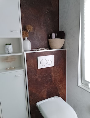 A bathroom with a white toilet, a white cabinet with a marble vase, a textured wall, and a window with blinds. - Beautiful Apartment in the Rhön (Kaltennordheim)
