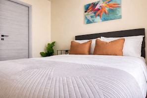 Un lit avec une couette blanche et deux oreillers marron, une porte grise et une plante verte.