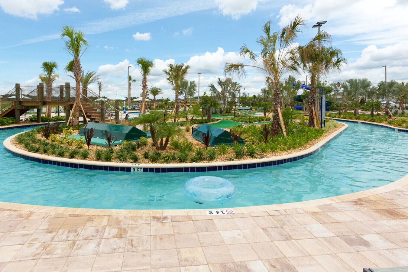 Une piscine dont la profondeur maximale est de 3 pieds, entourée de palmiers et traversée par un pont en bois.