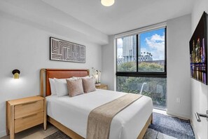 A bedroom with a bed, a nightstand, a wall-mounted artwork, a window with a city view, and a door. - Urban Corner 2 Bedrooms MIA, Wrap Balcony & Rooftop Pool (Miami)