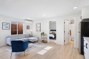 Ein modernes Wohnzimmer mit einer Couch, einem blauen Sessel, einem Couchtisch und einem Fernseher. Ein Flur führt zu einem Schlafzimmer mit einem Bett und einem Kleiderschrank.