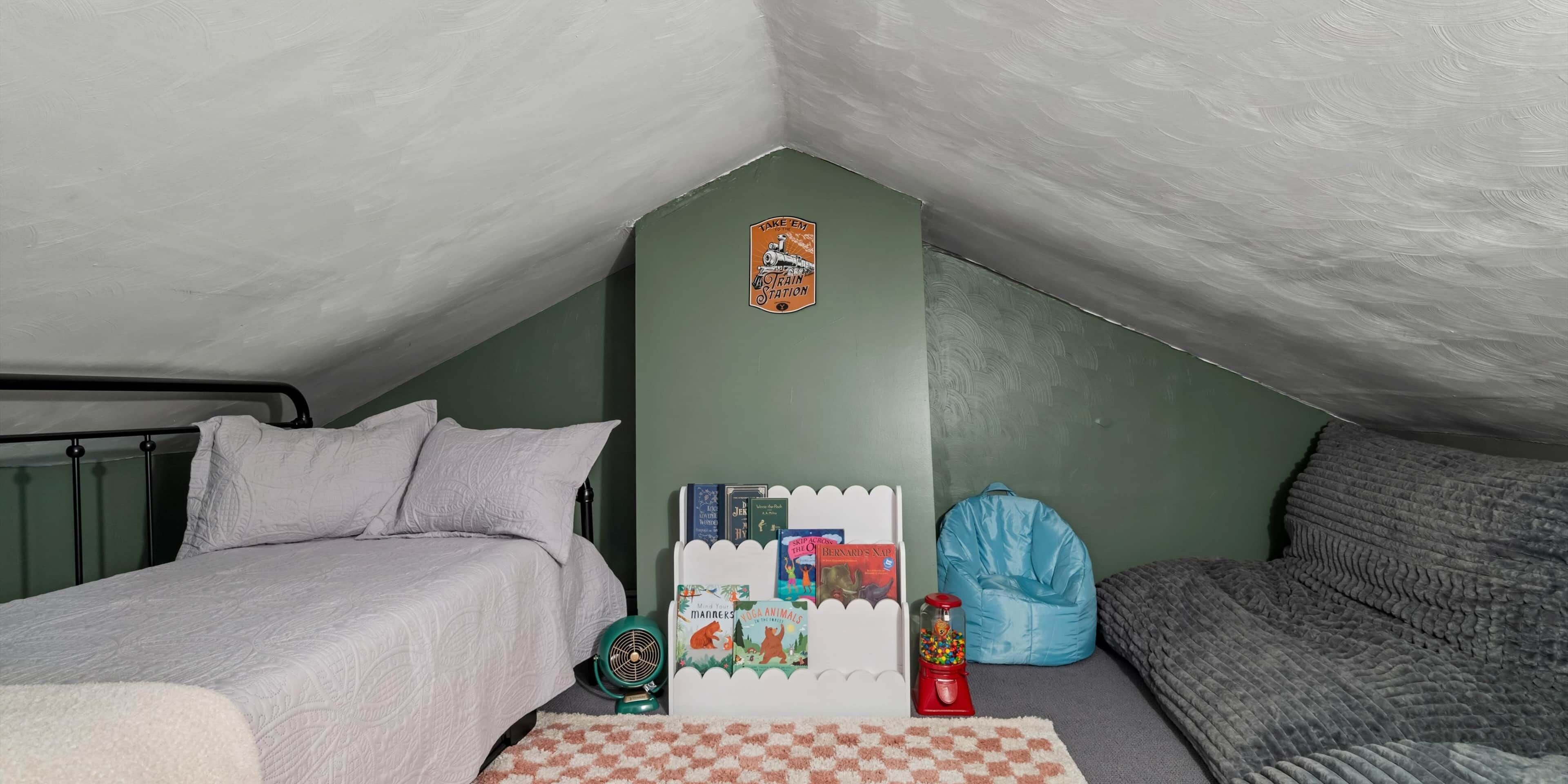 A small, cozy room with a sloped ceiling, a bed with white bedding, a green wall, a whiteboard, a bean bag, a small fan, and a bookcase with books.