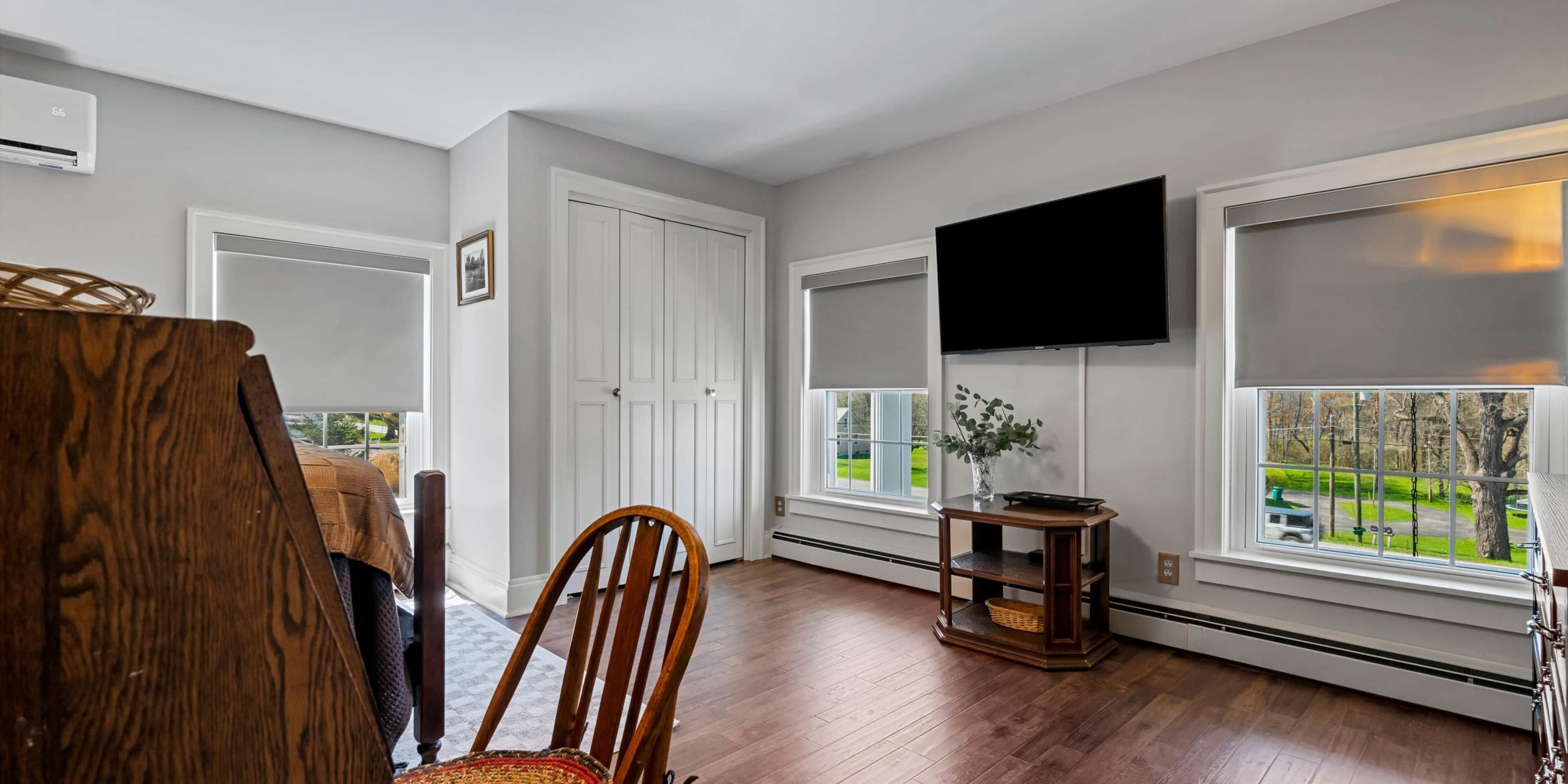 Una habitación con suelo de madera, una silla de madera, una puerta blanca, un televisor montado en la pared y una ventana con vistas a un paisaje verde.